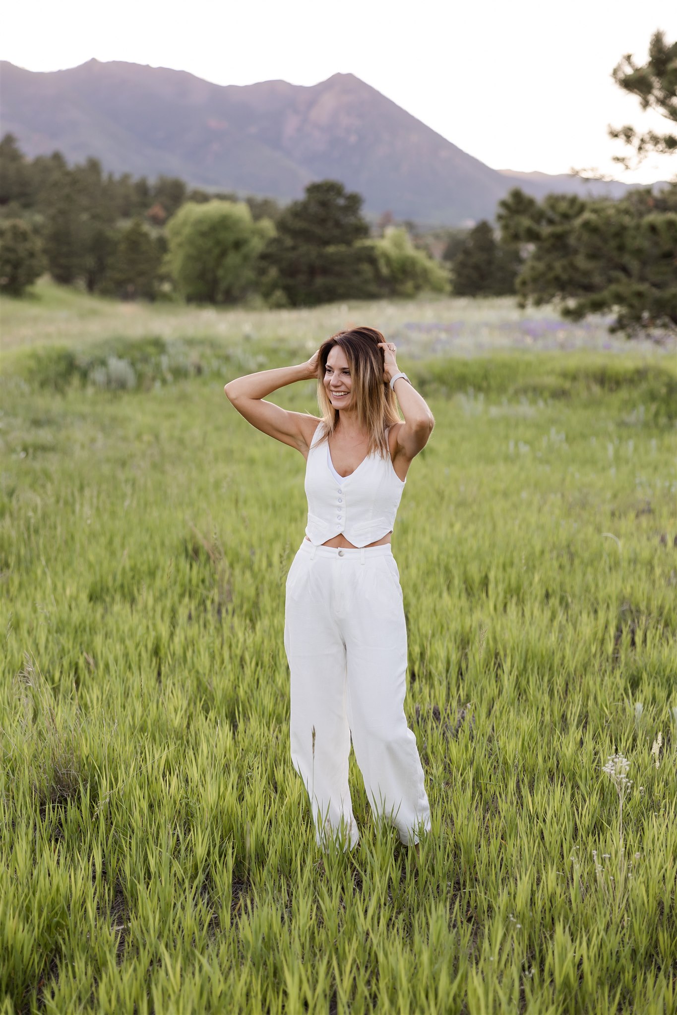 Female denver photographer poses with hands on her head in front of of Colorado mountains.