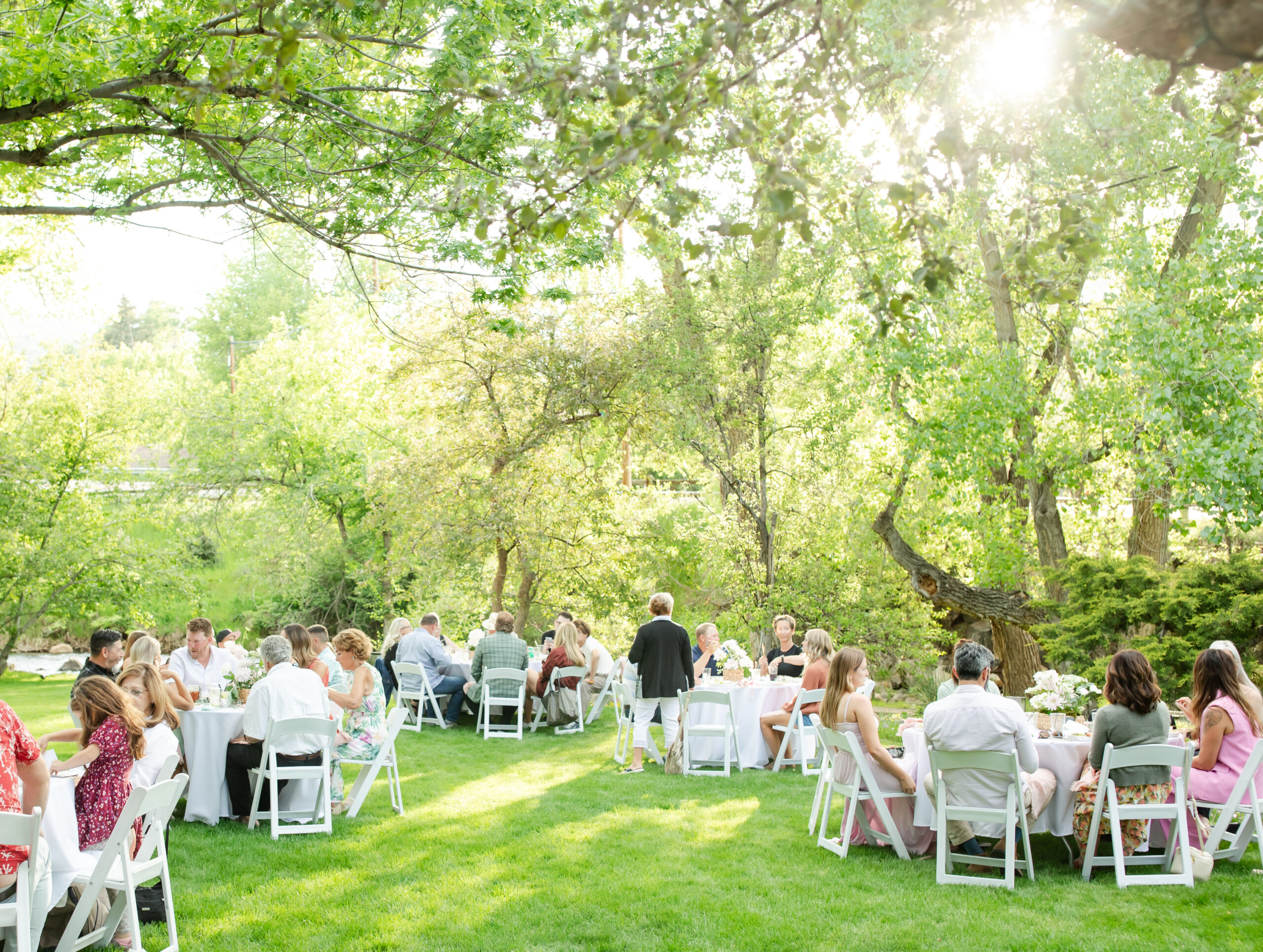 People sitting around linen covered tables in a garden area for a rehearsal dinner. Located in Morrison at a private residence.