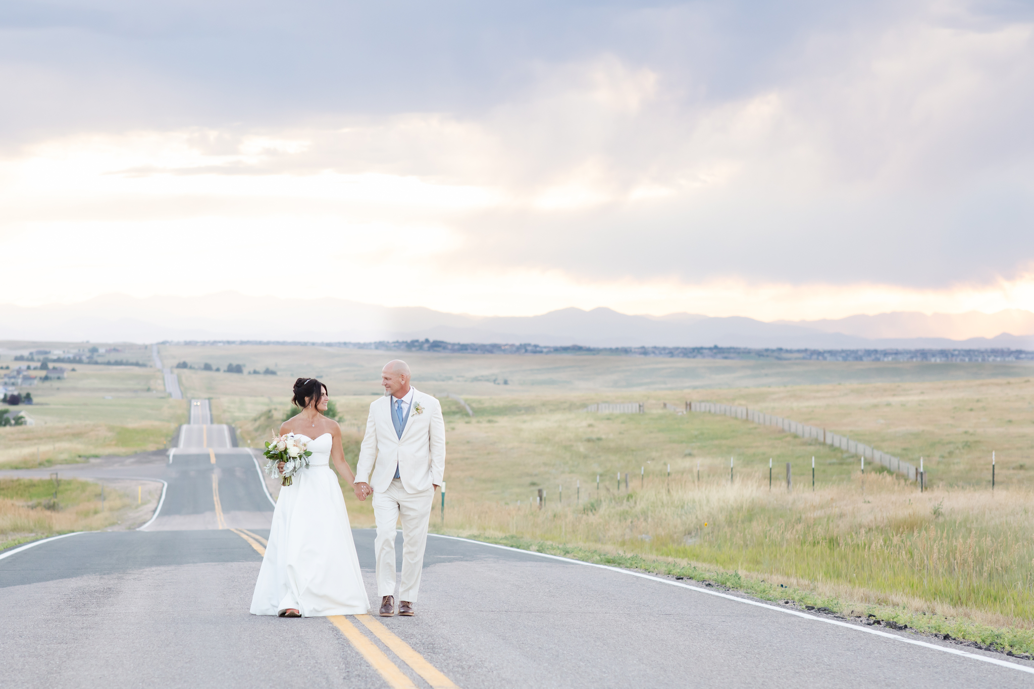 Bride and groom walk on blacktop road at sunset.
