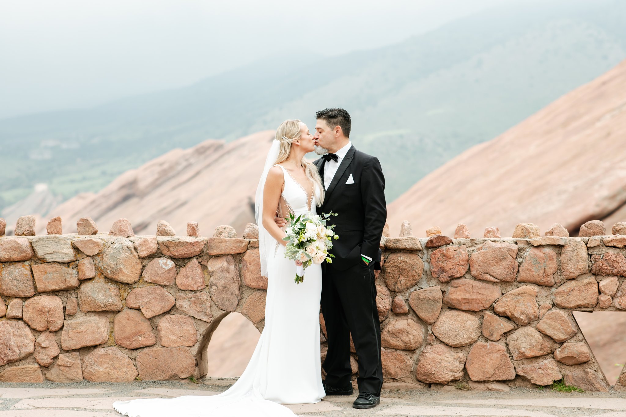 Happy newlyweds kiss on a patio with stone wall on mountain overlook