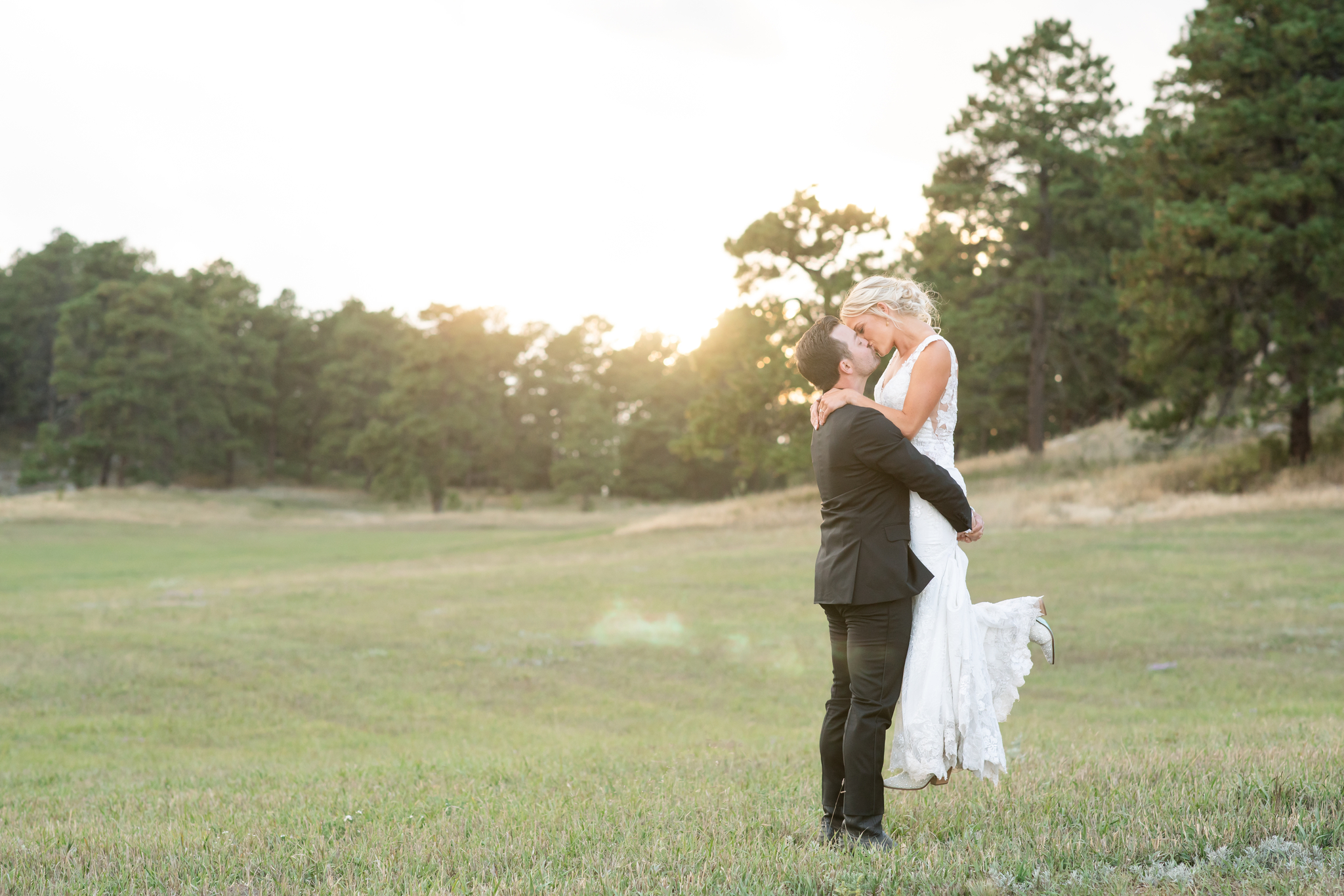 A groom lifts his bride for a kiss in a pasture at sunset