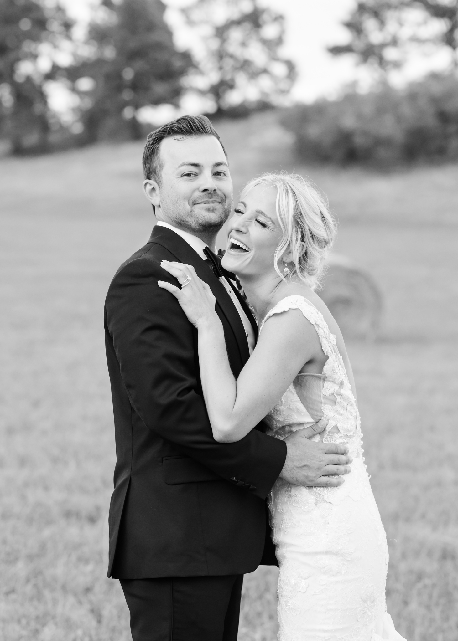 A bride laughs while hugging her groom in a tuxedo in a pasture at Wellshire Wedding Venue
