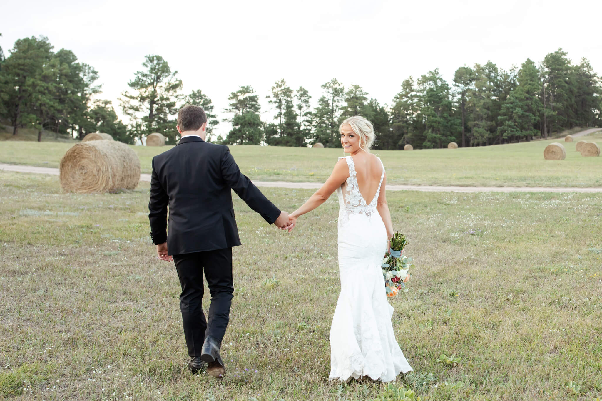 A bride smiles over her shoulder while walking in a hay field holding hands with groom thanks to first look events