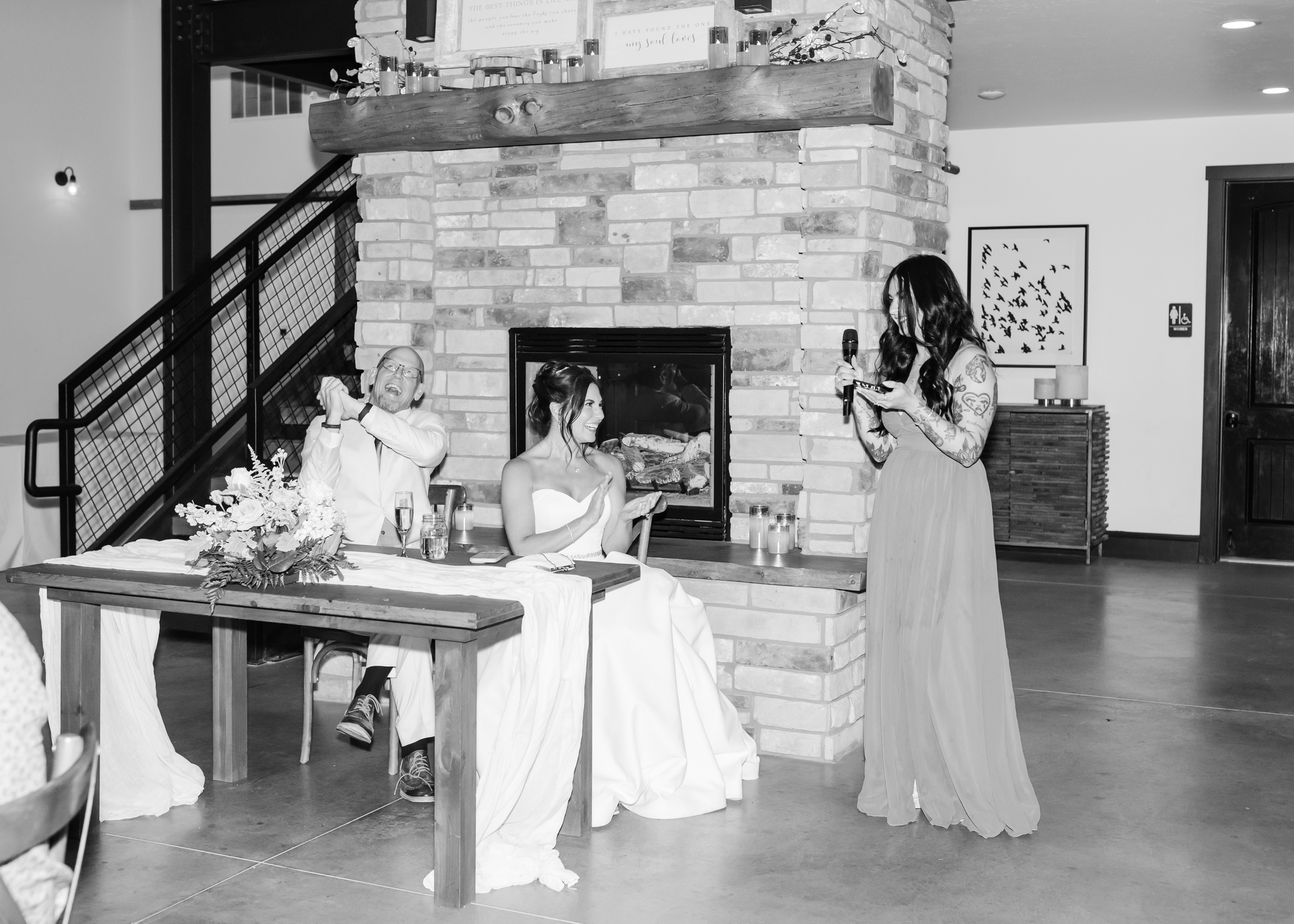 A bride and groom clap and laugh at their head table by fireplace as a bridesmaid gives a speech in a black and white photo