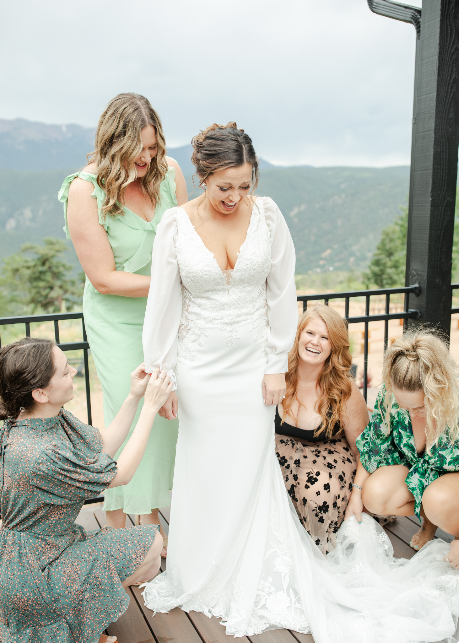 Bridesmaids laugh and admire the bride's gown and long train on a balcony in mountains